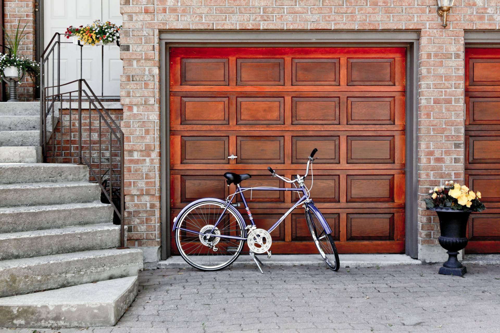 Vintage blue bicycle parked beside wooden garage doors and flower pot on a driveway, surrounded by brick walls.