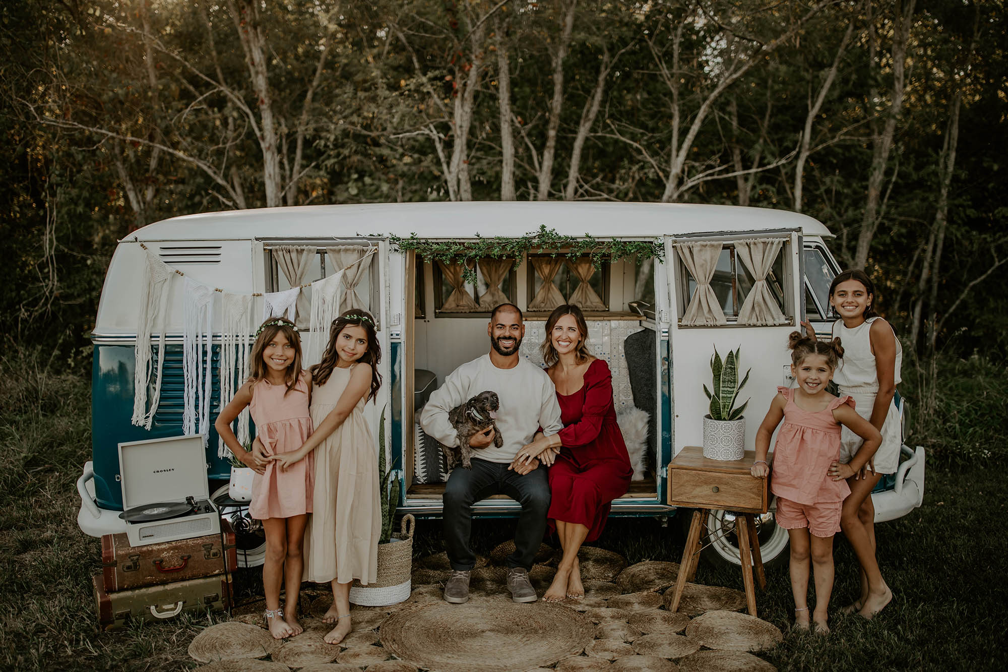 A joyful family poses by a vintage camper, surrounded by greenery, showcasing love and togetherness on a sunny day.