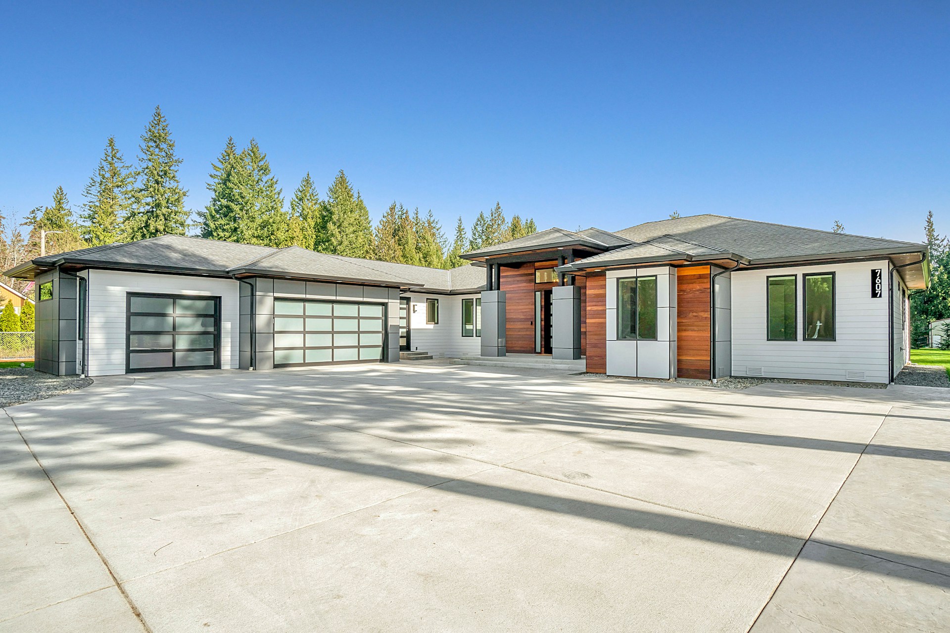 Modern single-story home with a two-car garage, set against a backdrop of lush green trees and clear blue sky.