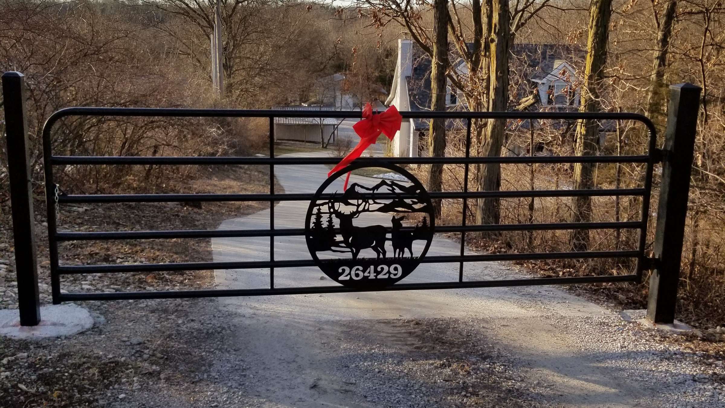 Black metal gate adorned with a red bow, leading to a gravel driveway surrounded by trees, featuring wildlife silhouette design.