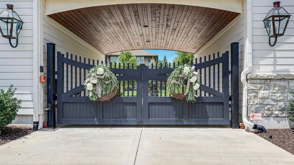 Elegant black gate adorned with floral arrangements, under a wooden archway, leading to a beautifully landscaped yard.