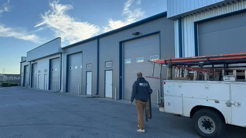 Man walking toward industrial storage units, with a truck loaded with equipment parked nearby under a blue sky.