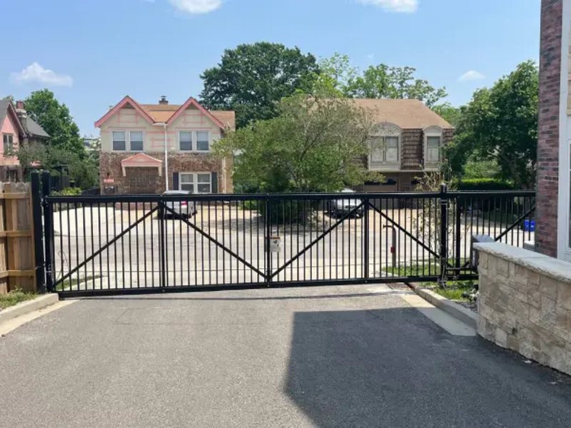 Black metal gate at driveway entrance, surrounded by residential homes and greenery under a clear blue sky.