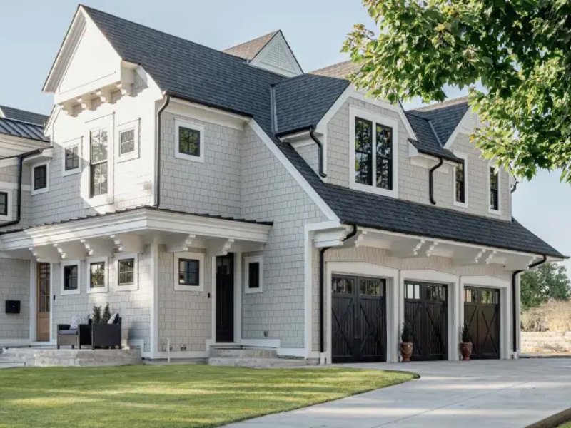 Beautiful modern home with gray shingles, porch, and three-car garage surrounded by lush greenery. Ideal for family living.