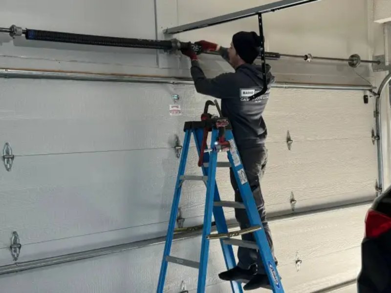 Technician on a ladder repairing a garage door opener mechanism in a residential garage.