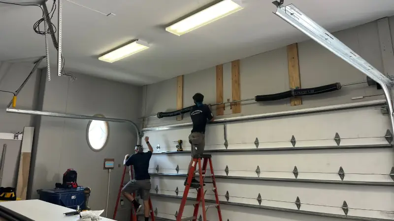 Two workers installing a garage door opener system on a wall with ladders in a well-lit garage setting.