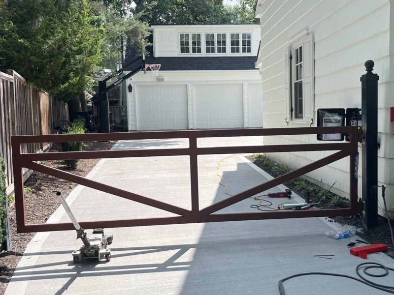 Brown metal driveway gate partially open at a residential property with a garage and driveway under bright sunlight.