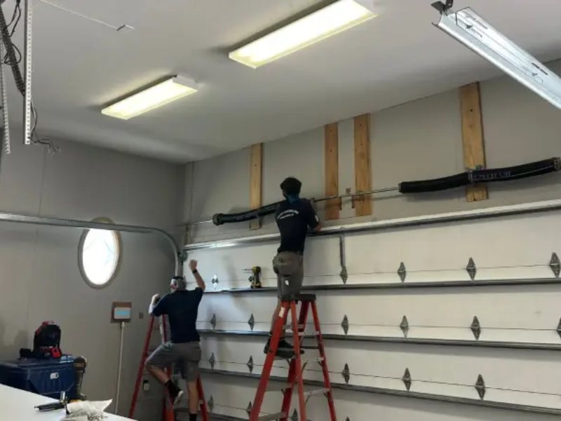 Two men on ladders installing a garage door opener in a spacious garage with overhead lighting.