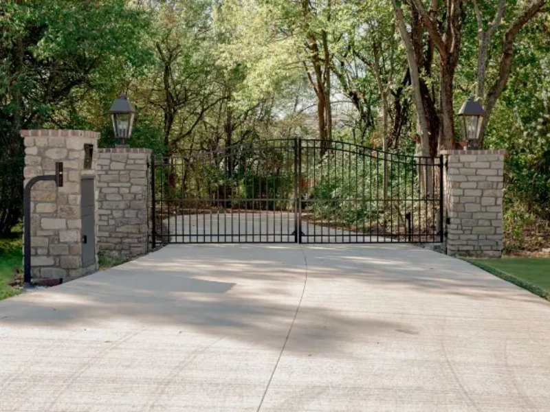 Elegant black wrought iron gate framed by stone pillars, leading to a lush, green landscape with trees.