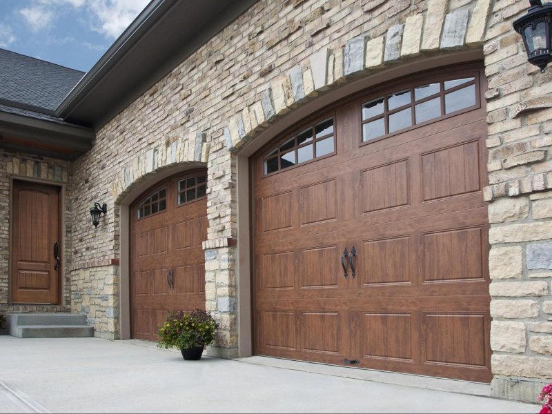 Elegant stone house entrance with two wooden garage doors and a potted plant on a concrete driveway.
