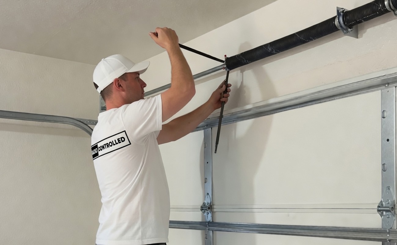 Man adjusting a garage door spring using a tool, wearing a Radio Controlled t-shirt in a light garage setting.
